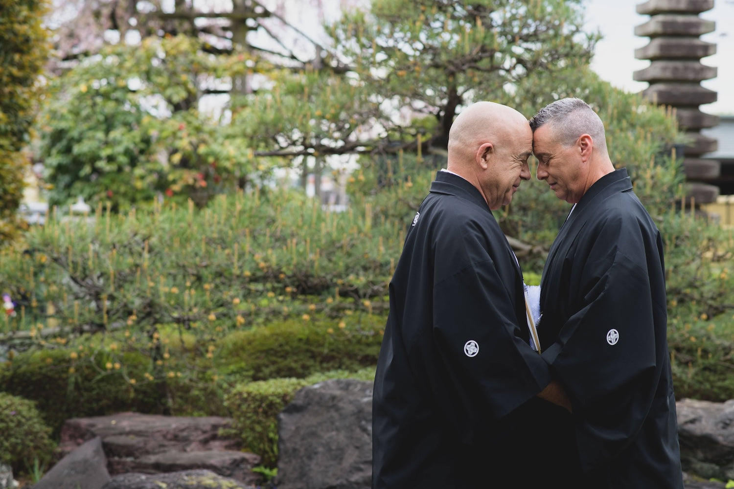 Two grooms embracing forehead to forehead in black montsuki hakama during a Shinto wedding ceremony in a Japanese garden